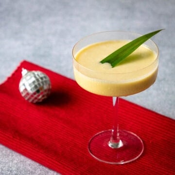 mango sticky rice cocktail in a coupe glass with a pandan leaf garnish and a small disco ball in the background.