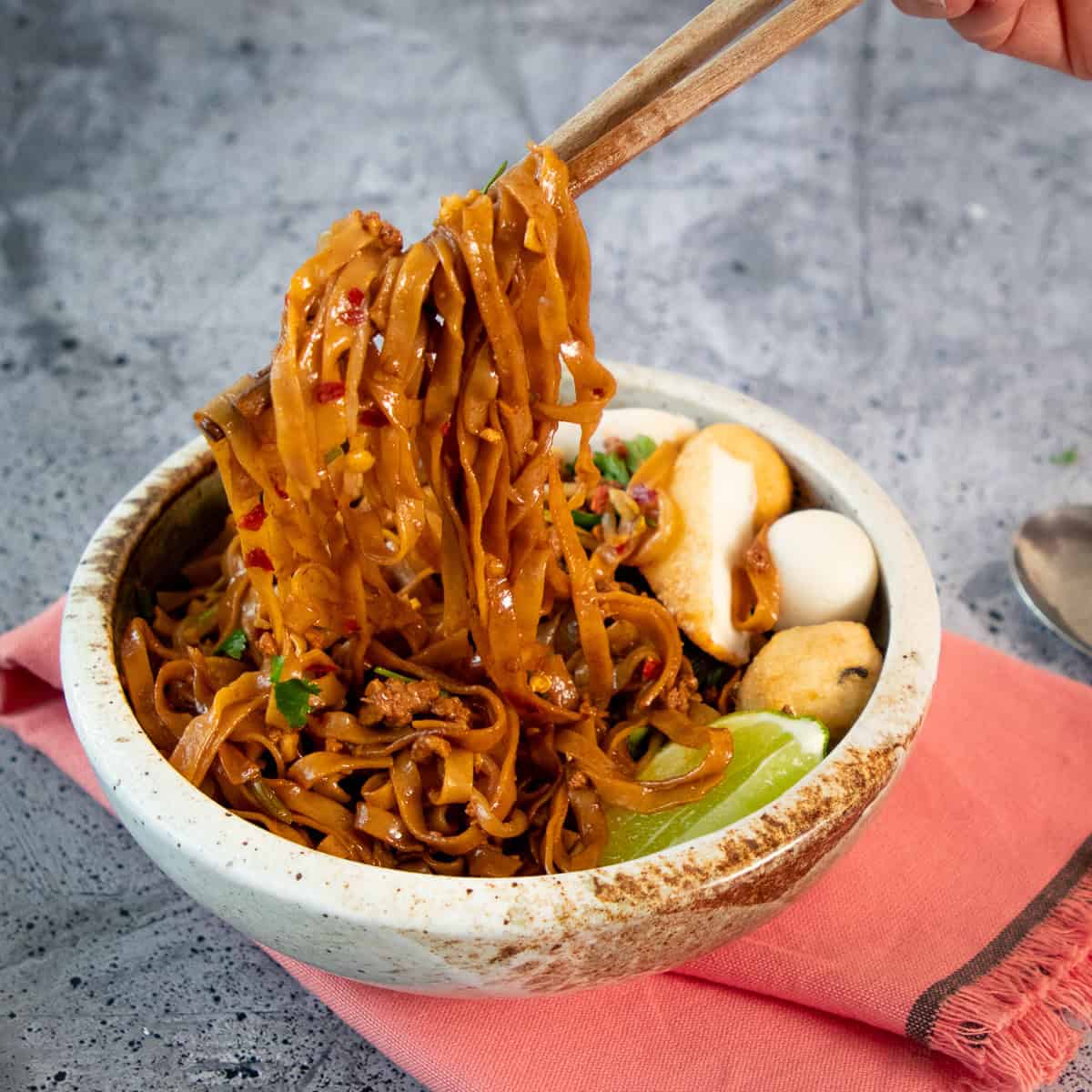 Chopsticks holding up tossed noodles in a bowl.